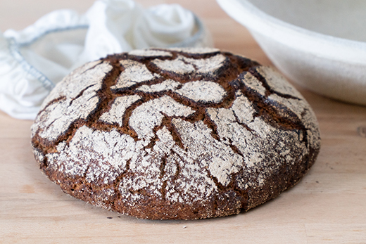 Rundes Sauerteigbrot mit dunkler Kruste und hellen Mehlrissen auf Holzuntergrund, im Hintergrund weiße Stoffabdeckung und Schüssel.