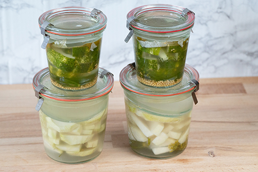 Vier Einmachgläser mit fermentiertem Gemüse, zwei mit grünen Gurken und Senfkörnern, zwei mit weißen Kohlrabi in Flüssigkeit.