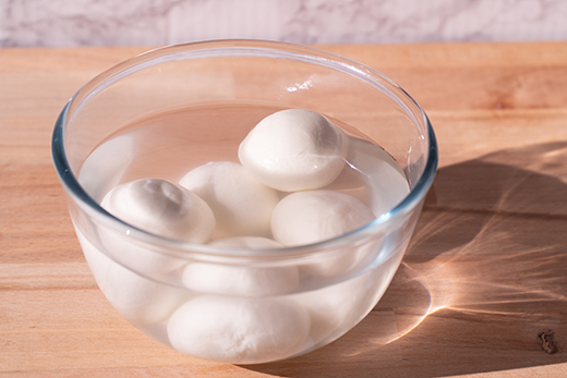 Glasschüssel mit mehreren weißen Mozzarellakugeln in Wasser auf Holzuntergrund.