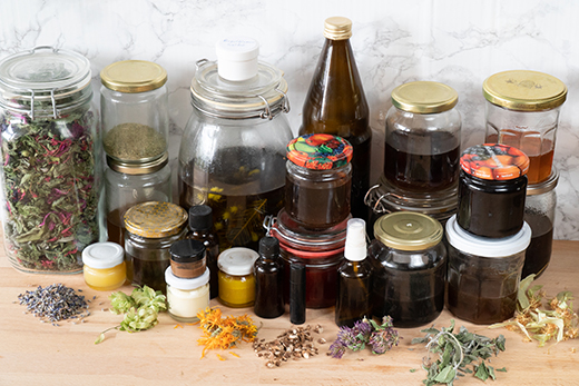 Verschiedene Glasgefäße mit getrockneten Kräutern, Ölen und Salben auf Holzuntergrund, davor getrocknete Blüten und Kräuter.