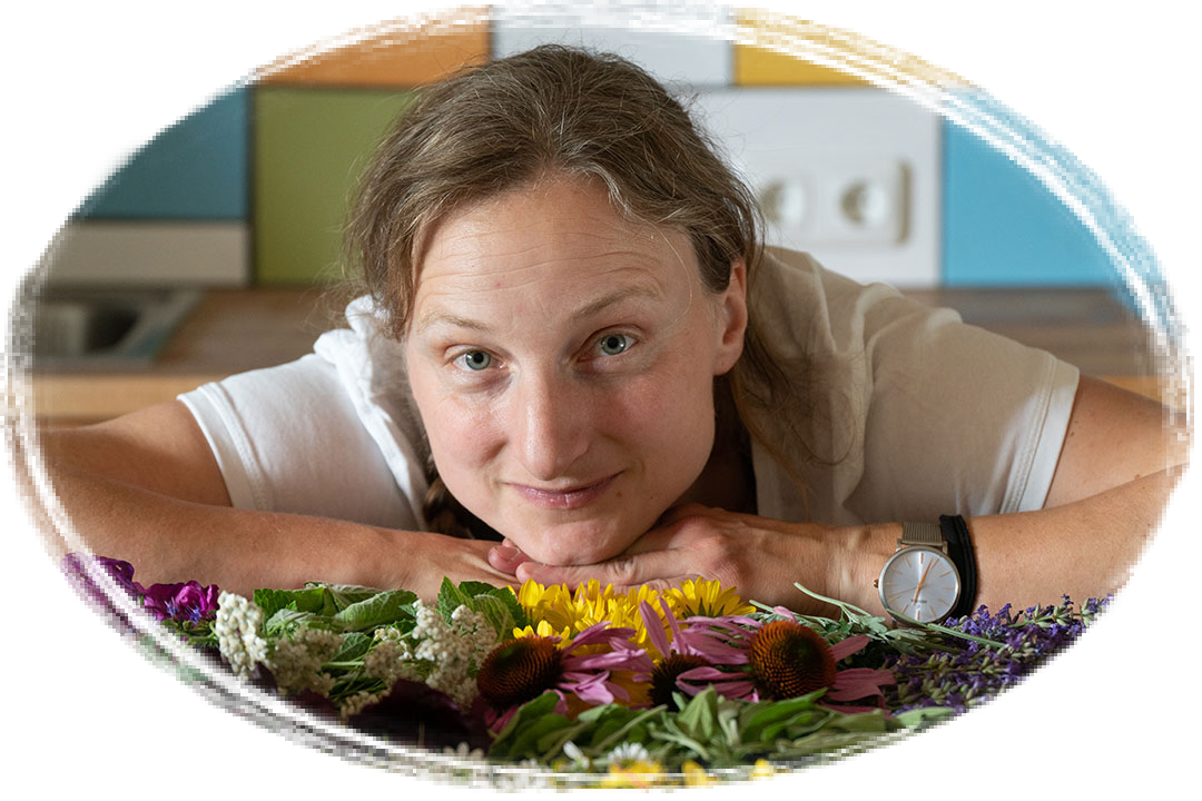 Frau mit weißem T-Shirt und Armbanduhr lehnt sich über einen Tisch mit verschiedenen bunten Kräutern und Blumen.