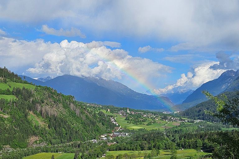 Regenbogen über grün bewaldeter Berglandschaft mit Wolken am Himmel.