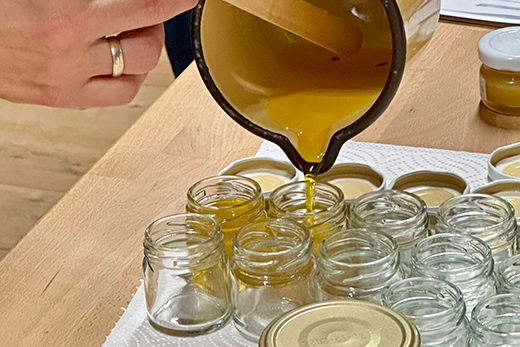 Hand gießt gelbliche Salbe aus einem kleinen Krug in mehrere kleine Glasgefäße auf einem Holztisch.
