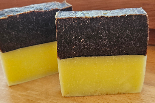 Zwei rechteckige Seifenstücke mit einer oberen dunkelbraunen und einer unteren gelben Schicht auf Holzuntergrund.