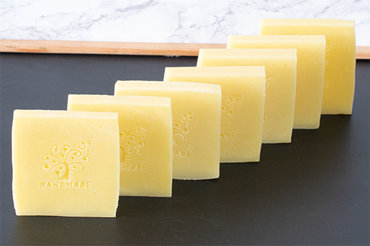 Sieben gelbliche, quadratische Seifenstücke mit Baum-Logo und Schriftzug 'HANDMADE' in einer Reihe auf schwarzer Oberfläche.