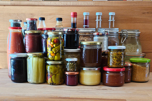 Verschiedene Gläser und Flaschen mit eingelegtem Gemüse, Soßen, Gewürzen und Honig auf einem Holzregal.