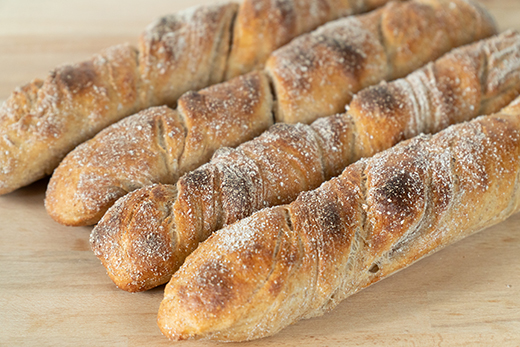 Vier längliche, goldbraun gebackene Wurzelbroten mit knuspriger Kruste und Mehlbestäubung auf Holzuntergrund.